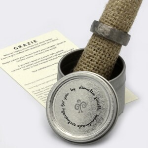 May include: A silver ring box with a burlap lining. The lid of the box has a silver label with the text "Exclusively for you by dimalta gioielli handmade" and a stylized swirl design.