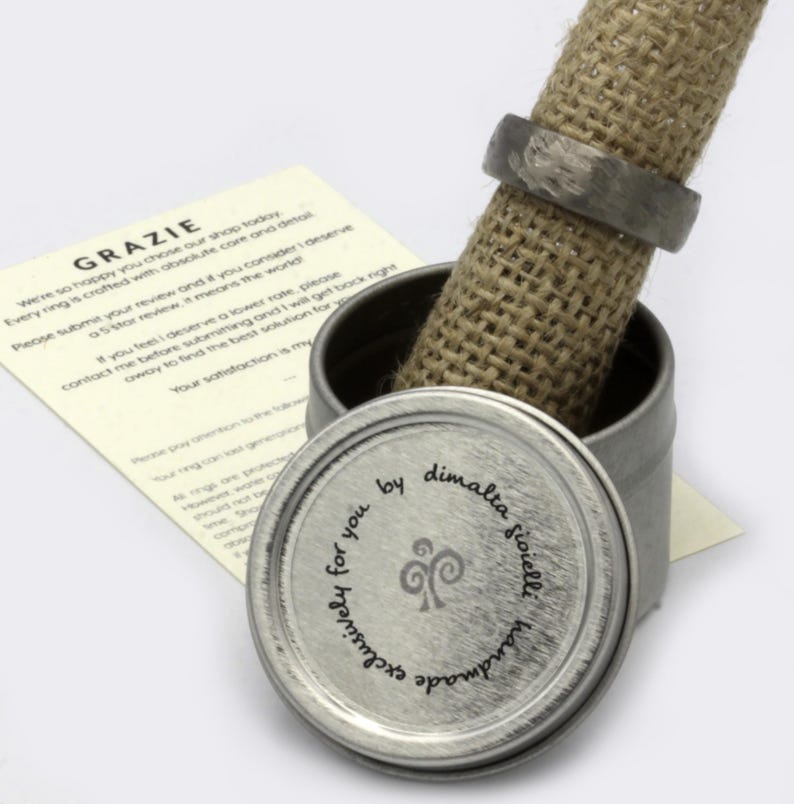 May include: A silver ring box with a burlap lining. The lid of the box has a silver label with the text "Exclusively for you by dimalta gioielli handmade" and a stylized swirl design.