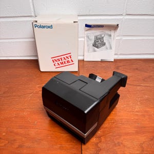 Peut inclure: Appareil photo instantané Polaroid noir avec une bordure argentée, posé sur une surface en bois. Une boîte blanche avec le logo Polaroid et le texte "INSTANT CAMERA" est derrière l'appareil photo, ainsi qu'un manuel.