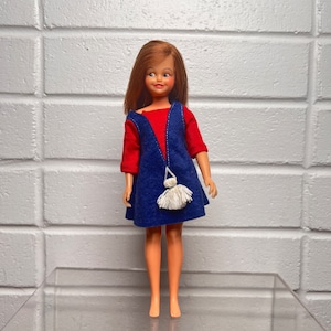 Peut inclure: Poupée vintage aux cheveux roux, vêtue d'un haut rouge et d'une robe bleue avec un sac à pompon blanc. La poupée est posée sur une surface transparente devant un mur de briques blanches.