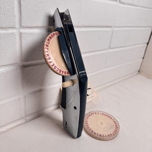 May include: A vintage Dymo M-20 label maker in silver and navy blue. The label maker has a beige dial with red letters and numbers. A spare dial is also visible. The label maker is on a white surface.
