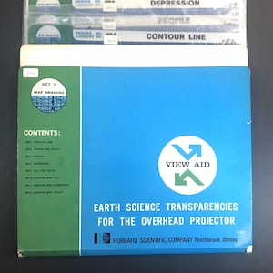 May include: A set of vintage Earth Science transparencies for an overhead projector. The blue and green box features the text "View Aid" and "Earth Science Transparencies." The contents include topics like contour lines, stream and valley, and depression.