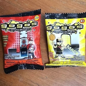 May include: Two colorful Gogo's Crazy Bones collectible packs. One is red with a character and a boombox graphic, the other is yellow with a basketball player graphic. Both are marked 4+ and contain small pieces.
