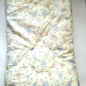 May include: A quilted baby blanket with a cream-colored background. The blanket is patterned with cartoon cats in various poses, wearing blue, pink, and purple outfits. Green leaf shapes are scattered throughout the design. A white zipper is visible along the bottom edge.