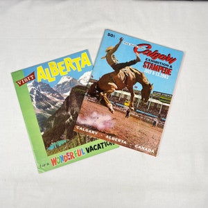 May include: Two vintage travel brochures. One green brochure reads "Visit Alberta" with a mountain landscape. The other, a Calgary Exhibition & Stampede souvenir program, features a cowboy on a bucking bronco, dated July 8-13, 1963.