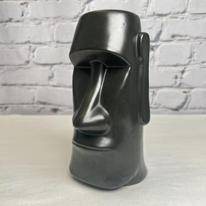 May include: A black ceramic mug shaped like a Moai statue from Easter Island. The mug has a large nose, prominent chin, and a simple, stylized design. The mug is approximately 6 inches tall.