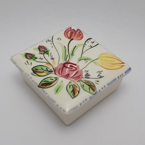 Puede incluir: Una caja de baratijas cuadrada de cerámica blanquecina con un diseño floral. La tapa presenta un arreglo pintado a mano de rosas rosas, hojas verdes y flores amarillas y rojas. La caja tiene un borde azul.