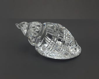 May include: A clear, faceted crystal figurine shaped like a seashell. The object features intricate, geometric cross-hatch cuts across its surface, creating a reflective, sparkling appearance that catches light from multiple angles.