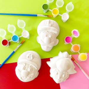May include: White plaster figures of Mario, Luigi, and Princess Peach, ready to be painted. Paint palettes with various colours and paintbrushes are arranged around the figures on a green and pink surface. A fun DIY craft project.