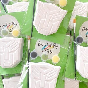 Peut inclure: Plusieurs kits de peinture à faire soi-même avec un motif d'emblème blanc, de petits pots de peinture gris, vert et jaune, et un pinceau. Les kits sont emballés dans du plastique transparent avec un support vert. Le texte "Brush & Play Artists" est visible.