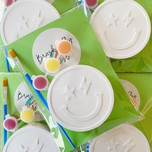 May include: White paint-your-own smiley face kits with star and lightning bolt designs. Each kit includes a paint palette with various colours and a paintbrush. The kits are packaged in clear plastic bags with green backing and the text "Brush my day artists".