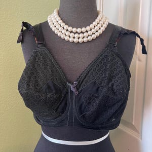 May include: Black lace bra with a black satin center panel and adjustable straps. The bra is on a mannequin wearing a pearl necklace.