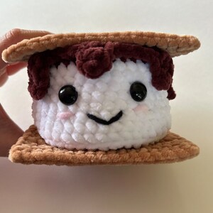 May include: A crocheted plush toy shaped like a s'more. The marshmallow is white with a brown chocolate topping and a smiling face. The graham crackers are brown.