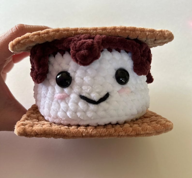 May include: A crocheted plush toy shaped like a s'more. The marshmallow is white with a brown chocolate topping and a smiling face. The graham crackers are brown.