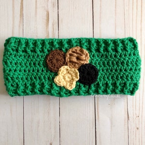 May include: Green crocheted headband with a brown, tan, and black flower design. The headband is made with a textured stitch and has a small tag that says "CrochetbyClaudia".