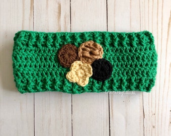 May include: Green crocheted headband with a brown, tan, and black flower design. The headband is made with a textured stitch and has a small tag that says "CrochetbyClaudia".