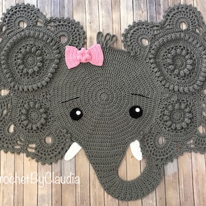 May include: A gray crocheted elephant head with large, detailed ears. The elephant has black eyes, white tusks, and a pink bow. The design is handmade and suitable for nursery decor.