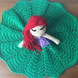 May include: A handmade crochet doll with red hair and a purple top, resting on a large, scalloped green crochet blanket. The doll has closed eyes and white arms. The blanket has a textured pattern.