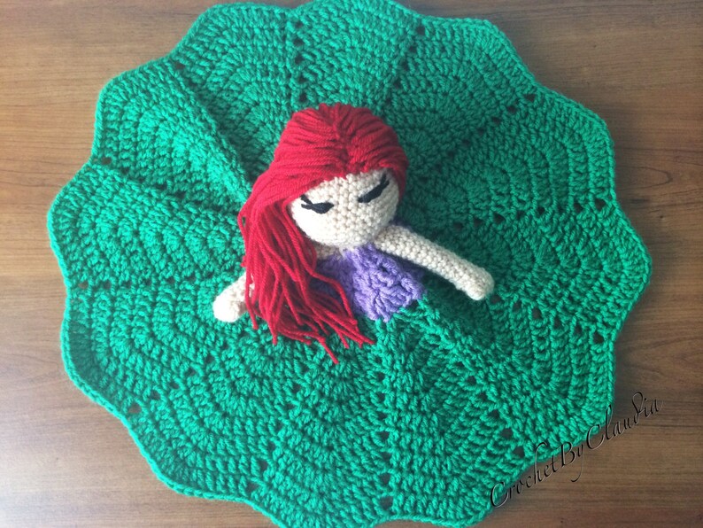 May include: A handmade crochet doll with red hair and a purple top, resting on a large, scalloped green crochet blanket. The doll has closed eyes and white arms. The blanket has a textured pattern.