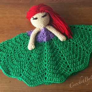 May include: A handmade crochet doll with red hair, a beige face, purple top, and green skirt. The doll is lying on a wooden surface. The doll's skirt has a scalloped edge.