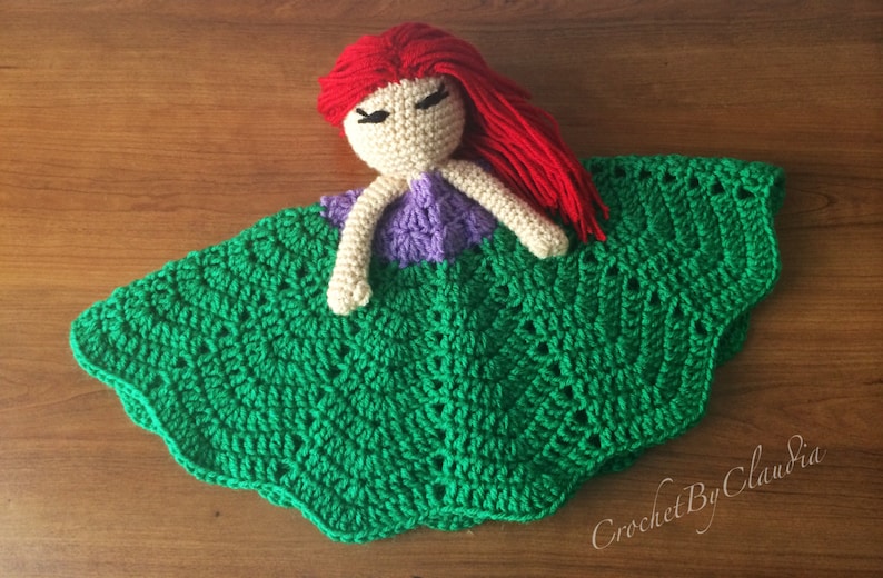May include: A handmade crochet doll with red hair, a beige face, purple top, and green skirt. The doll is lying on a wooden surface. The doll's skirt has a scalloped edge.