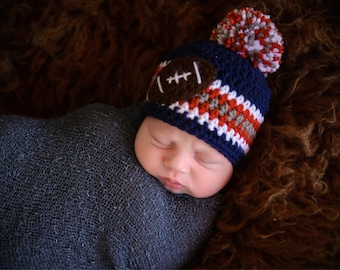 May include: A baby wearing a navy blue, white, red, and grey crocheted football-themed hat with a brown football applique and a matching pom-pom. The baby is wrapped in a dark blue blanket, resting on a brown, textured surface.