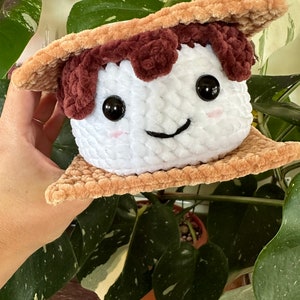 May include: A plush toy shaped like a s'more. The marshmallow is white with brown eyes and a smiling mouth. The graham crackers are brown and textured.