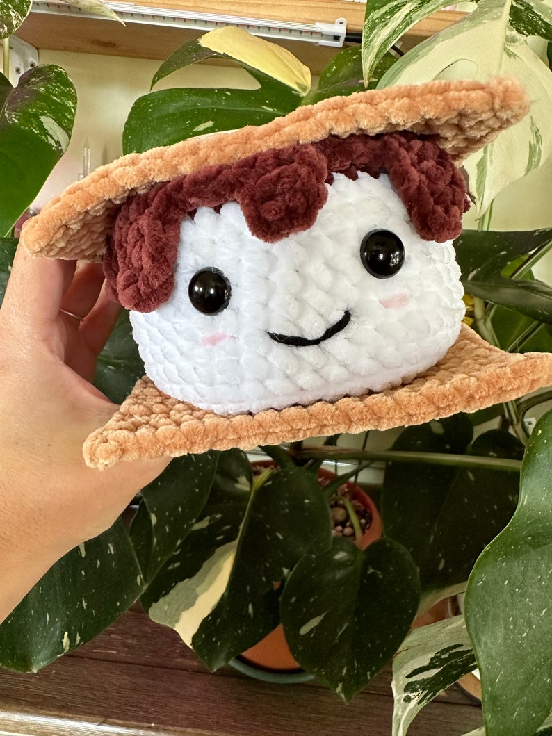 May include: A plush toy shaped like a s'more. The marshmallow is white with brown eyes and a smiling mouth. The graham crackers are brown and textured.