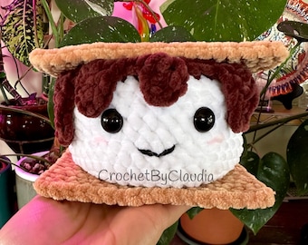 Könnte beinhalten: Ein gehäkeltes Plüschtier in Form eines Marshmallow-Brötchens. Das Marshmallow ist weiß mit brauner Schokolade und einem lächelnden Gesicht. Die Graham-Cracker sind braun. Das Spielzeug ist mit "CrochetByClaudia" beschriftet.