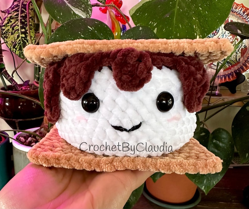 May include: A crocheted plush toy shaped like a s'more. The marshmallow is white with brown chocolate and a smiling face. The graham crackers are brown. The toy is labeled "CrochetByClaudia".