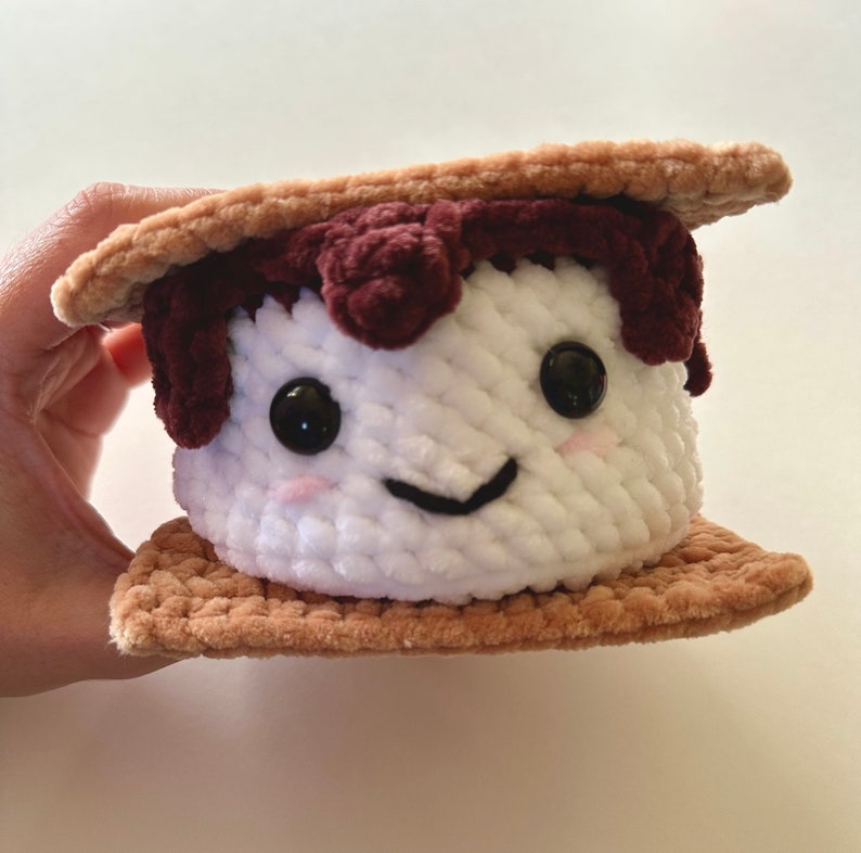 May include: A crocheted plush toy shaped like a s'more. The marshmallow is white with a smiling face and brown chocolate on top. The graham crackers are brown.