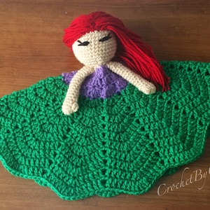 May include: Crocheted doll with red hair, a beige face, and a purple top, resting on a green crocheted blanket. The blanket has a scalloped edge and a textured pattern. The doll's hair is made of red yarn.