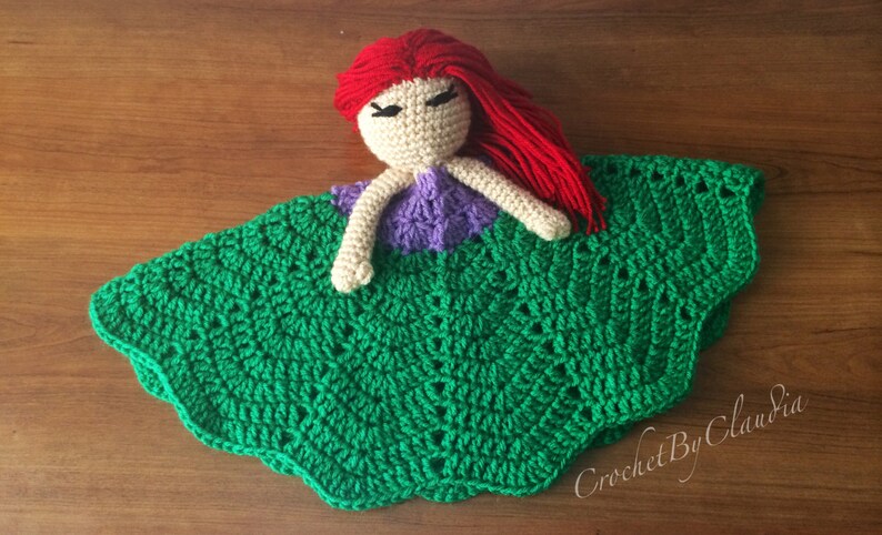 May include: Crocheted doll with red hair, a beige face, and a purple top, resting on a green crocheted blanket. The blanket has a scalloped edge and a textured pattern. The doll's hair is made of red yarn.