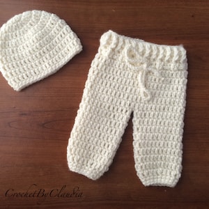 May include: A cream-colored, crocheted baby set, including a beanie hat and pants with a drawstring. The set is handmade and displayed on a brown wooden surface. The text "Crochet By Claudia" is visible in the bottom left corner.