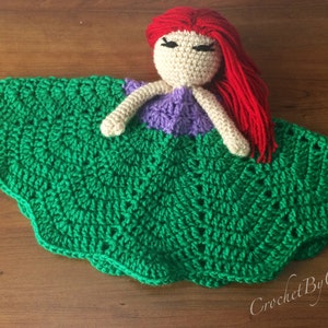 May include: A handmade crochet doll with a red-haired head, beige face, and purple top, resting on a green, scalloped blanket. The doll's arms are beige. The blanket has a detailed, textured pattern. The doll is a handmade item.
