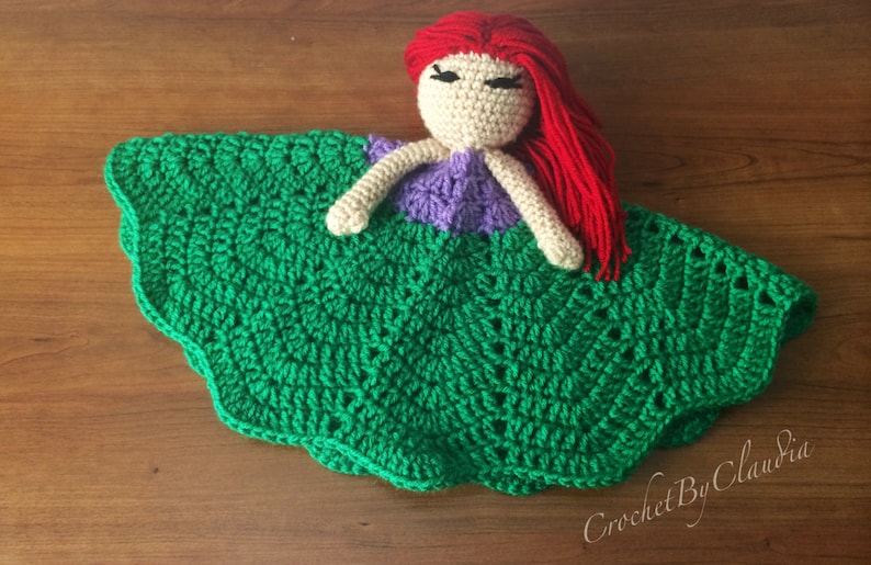 May include: A handmade crochet doll with a red-haired head, beige face, and purple top, resting on a green, scalloped blanket. The doll's arms are beige. The blanket has a detailed, textured pattern. The doll is a handmade item.