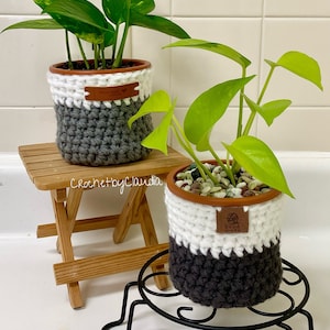 May include: Two potted plants with green leaves sit on a small wooden table and a black metal plant stand. The pots are wrapped in a crochet design with white, gray, and brown yarn. Each pot has a brown leather tag that says "Hand Made".