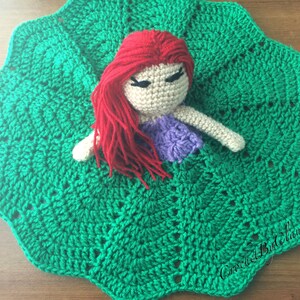 May include: A handmade crochet doll with red hair, a purple top, and beige skin rests on a green crochet blanket. The blanket has a scalloped edge and a textured pattern. The doll's eyes are closed. The overall theme is handmade crafts.