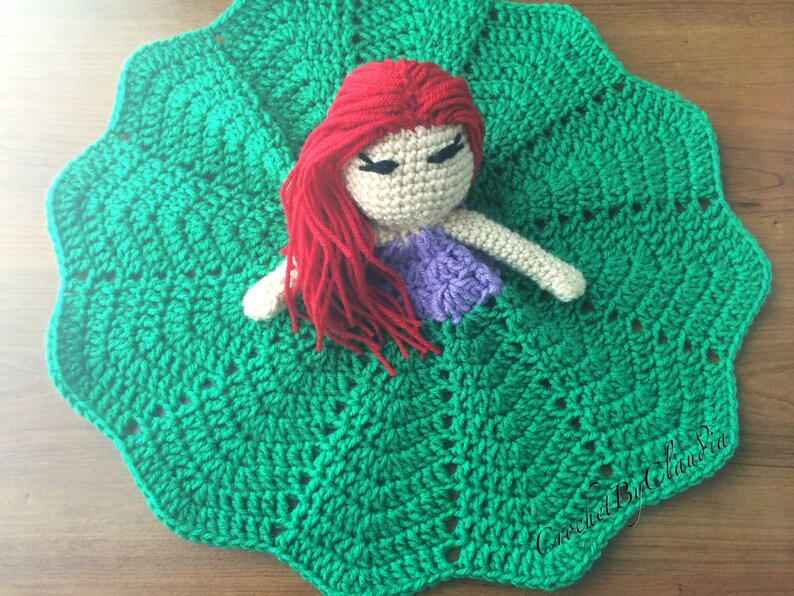 May include: A handmade crochet doll with red hair, a purple top, and beige skin rests on a green crochet blanket. The blanket has a scalloped edge and a textured pattern. The doll's eyes are closed. The overall theme is handmade crafts.