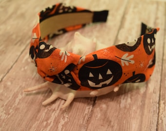 May include: Orange headband with a black and white Halloween pumpkin pattern. The headband is knotted in the center.