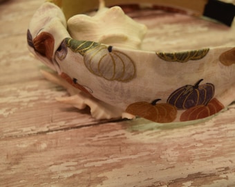 May include: A white headband with a knotted design and a fall-themed print of pumpkins in shades of orange, purple, and green.