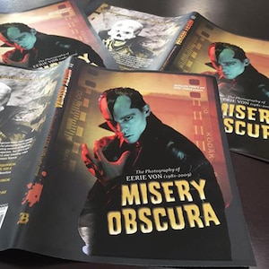 May include: A black book cover with a title that reads "The Photography of Eerie Von (1981-2009) Misery Obscura". The cover features a close-up of a person with blue skin and a black and red outfit.