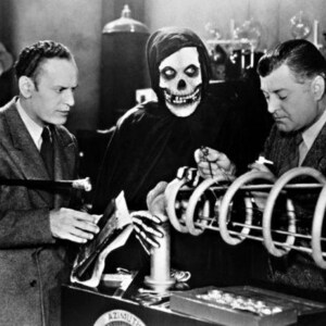 May include: A black and white photo of three people in a laboratory setting. One person is wearing a hooded robe with a skull mask. The other two people are looking at a large, cylindrical device.