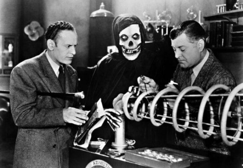 May include: A black and white photo of three people in a laboratory setting. One person is wearing a hooded robe with a skull mask. The other two people are looking at a large, cylindrical device.