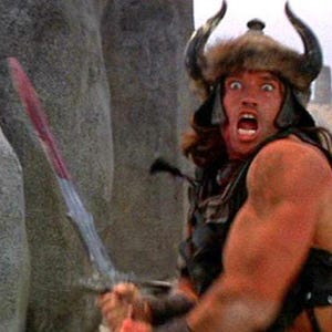 May include: A man wearing a horned helmet and fur-lined cape holds a sword with a red handle. He has a shocked expression on his face. The background is a desert landscape with large rocks.