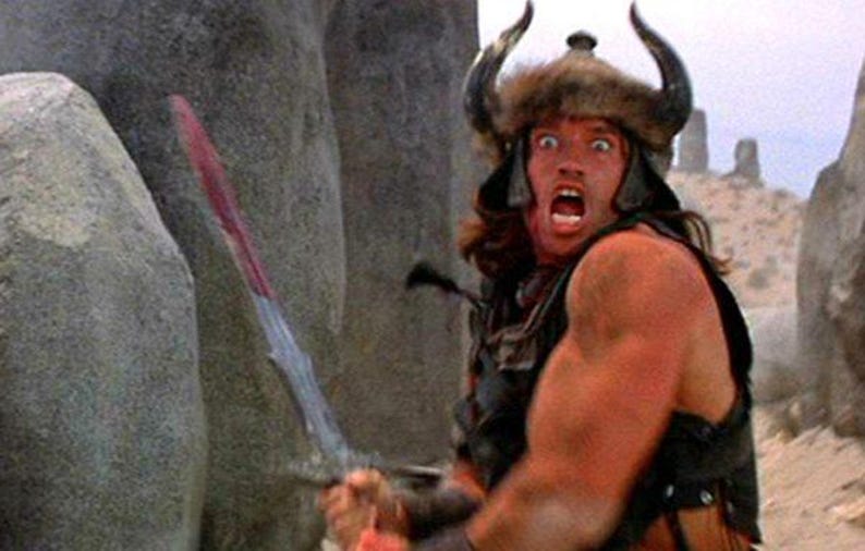 May include: A man wearing a horned helmet and fur-lined cape holds a sword with a red handle. He has a shocked expression on his face. The background is a desert landscape with large rocks.