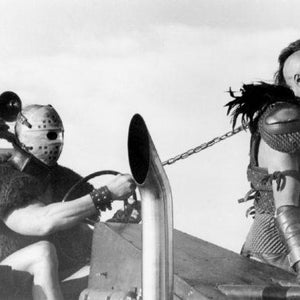 May include: A black and white photo of two men in a post-apocalyptic setting. One man is wearing a helmet and a chainmail shirt, while the other man is wearing a leather jacket and a helmet. They are both holding weapons and appear to be in a fight.