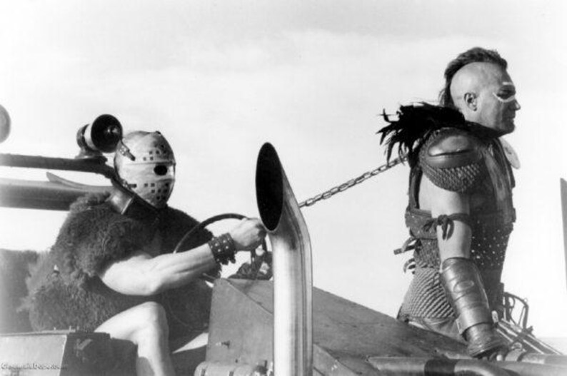 May include: A black and white photo of two men in a post-apocalyptic setting. One man is wearing a helmet and a chainmail shirt, while the other man is wearing a leather jacket and a helmet. They are both holding weapons and appear to be in a fight.