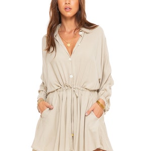 Puede incluir: Un vestido camisa beige de manga larga con cierre de botones, cintura con cordón y dos bolsillos. El vestido tiene un corte holgado y una falda ligeramente acampanada.