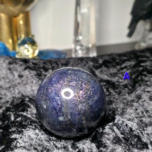 May include: A dark blue crystal sphere with shimmering inclusions. The sphere is set against a black velvet surface. Other objects are visible in the background, including a gold-colored decorative item and a clear glass bottle.